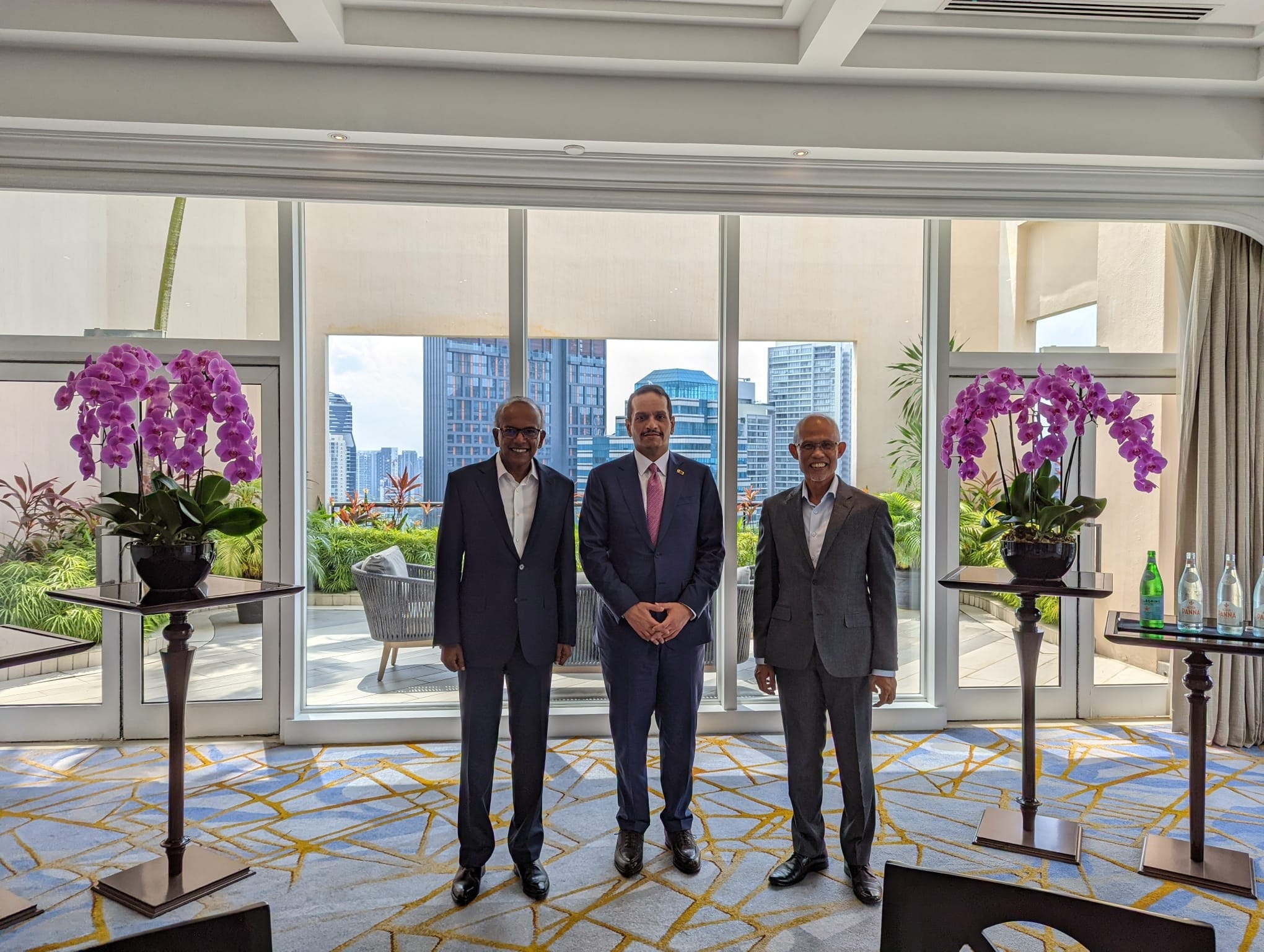 Three men in suits stand inside, flanked by orchids; city buildings visible through the window wall.